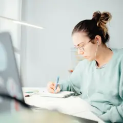 Student med hörlurar skriver i anteckningsbok vid skrivbordet, inspirerad av sitt universitetsutbildning i kreativt skrivande.