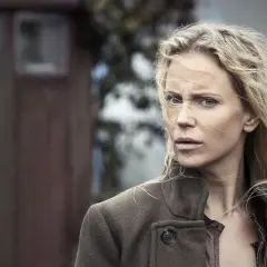 En kvinna med blont hår, klädd i en brun jacka, tittar oroligt in i kameran. Bakgrunden är suddig och antyder en utomhusmiljö, kanske från en bron serie.