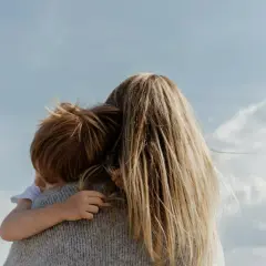 En mamma kramar sitt barn under en blå himmel med moln. En varm kram mellan mammor och minimodeller.