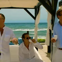 Tre män i vita och ljusblå skjortor dricker ur champagneglas vid havet. En scen från en exit serie.