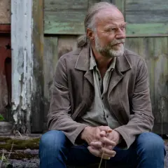 Hans Gunnarsson sitter utanför ett gammalt, slitet hus med en grön dörr och röd snickarglädje.