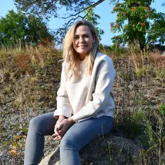 Ulrika Rolfsdotter sitter på en sten utomhus, omgiven av gräs och träd.