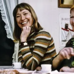 Två skrattande kvinnor vid ett bord. En av dem, med randig tröja, är en av skådespelarna från "Hem till byn".