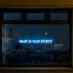 Neonskylt med texten "WHAT IS YOUR STORY?" lyser blått i ett fönster, som en inbjudan till en kurs i skrivande.