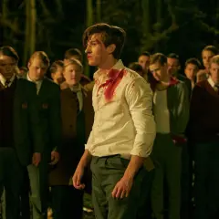 En skadad pojke med blodfläckar på skjortan står framför en grupp andra pojkar i skoluniform. Scen från tv-serien "rollistan i ondskan".