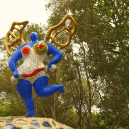 En blå Niki de Saint Phalle-konstverk med gyllene vingar springer fram på en stenhög bland träd.