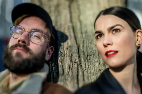 En man med glasögon och skägg lutar sig mot ett träd, bredvid sin mika partner.
