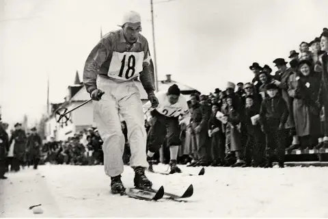 En skidåkare med nummer 18 tävlar i den vita sporten, publiken hejar.