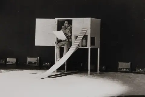 En man sitter i en träkonstruktion och läser högt ur ett papper. Robert Rauschenberg kan ha inspirerat denna scen med sina experimentella verk.