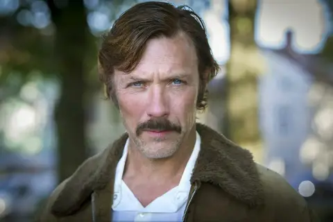 En manlig svensk skådespelare med blå ögon och mustasch, klädd i en brun jacka med foder.