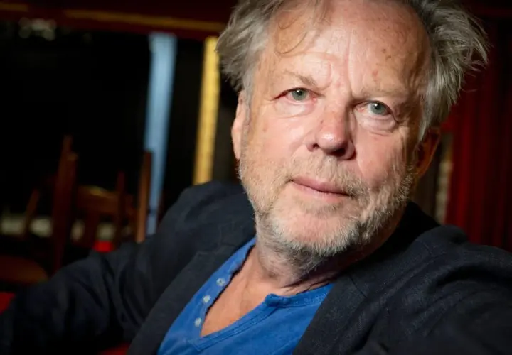 En äldre man med grått hår och skägg, klädd i kavaj och blå tröja. Han påminner om en ung Wallander.