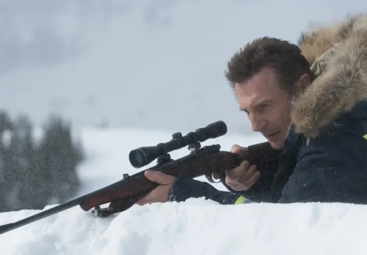 Liam Neeson i en vinterjacka med luva siktar med ett gevär i snön.