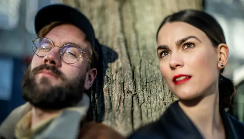 En man med glasögon och skägg lutar sig mot ett träd, bredvid sin mika partner.