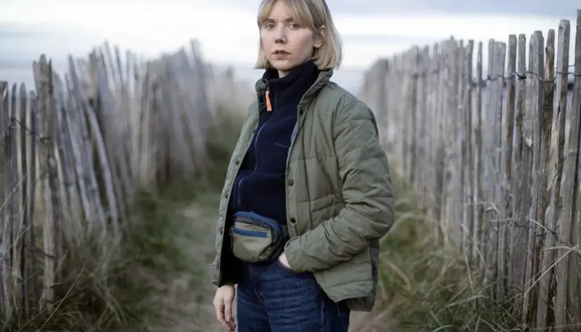 Karen Pirie står vid en vindpinad strand, klädd i en grön jacka och jeans. Hon bär en midjeväska, redo för ett nytt fall.