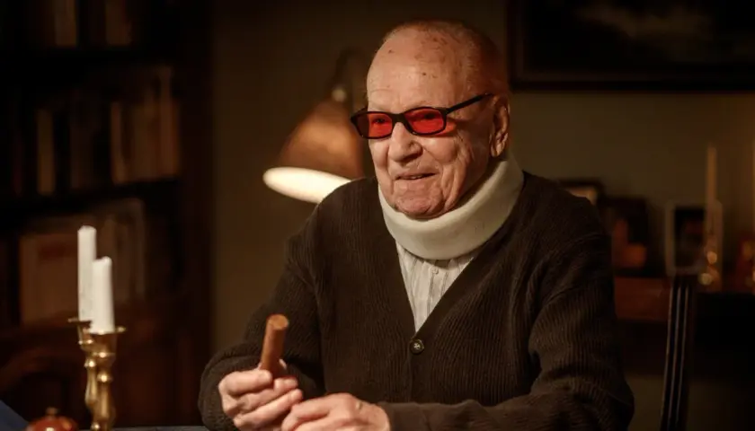 Äldre man med nackkrage och röda glasögon håller en cigarr. Han ser ut som grannen i beck, redo för en stunds avkoppling.