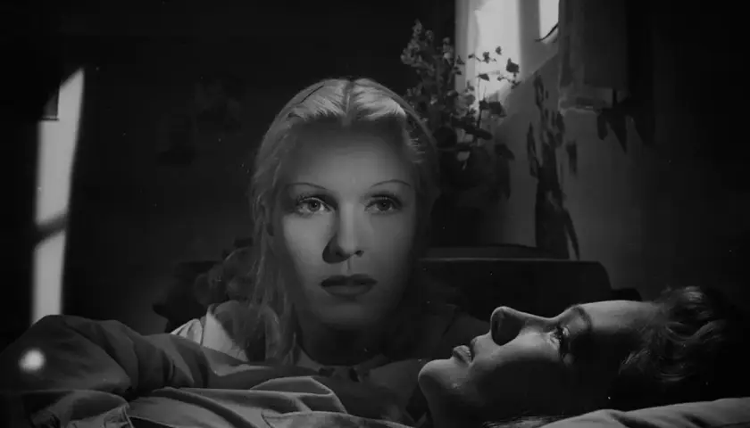 De två kvinnorna i dramatisk belysning. En kvinna ligger ner, medan den andra tittar ner på henne med intensiv blick.