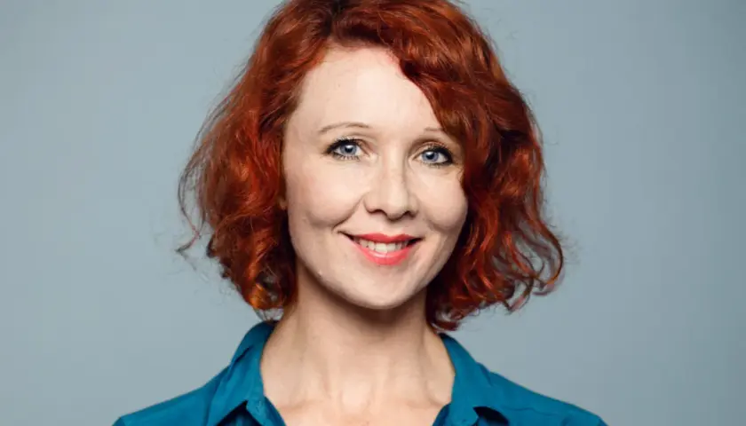 Ellinor Skagegård, en kvinna med rött lockigt hår och blå ögon, ler mot kameran i en blå skjorta.