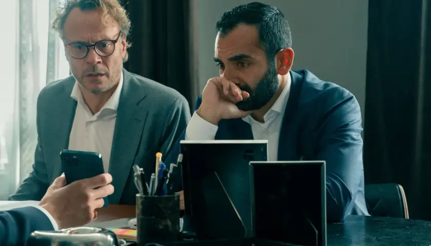Två män i kostym sitter vid ett bord. Den ena mannen, med glasögon, tittar på sin mobiltelefon. Den andra mannen, med skägg, funderar djupt. Scen från "hundarna" på SVT.