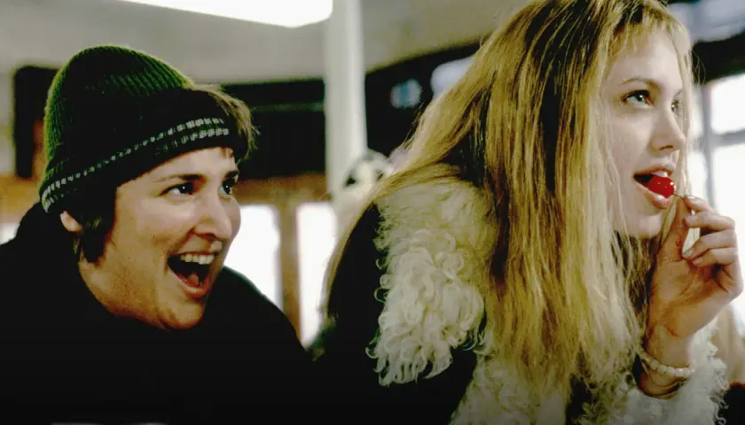 En glad kvinna med grön mössa och en annan kvinna med blont hår som äter ett körsbär. Scen från "Girl, Interrupted" (Stulna år).