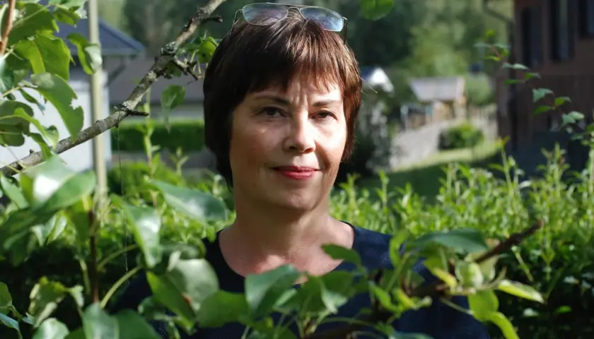 Anna Bratt, en kvinna med glasögon på huvudet, ler mot kameran i en lummig trädgård.