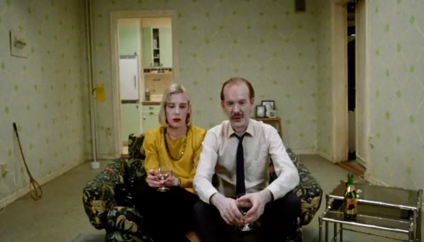 Ett par sitter i en soffa, en kvinna med gul tröja och en man i skjorta och slips. Kanske en scen från en Roy Andersson reklamfilm.