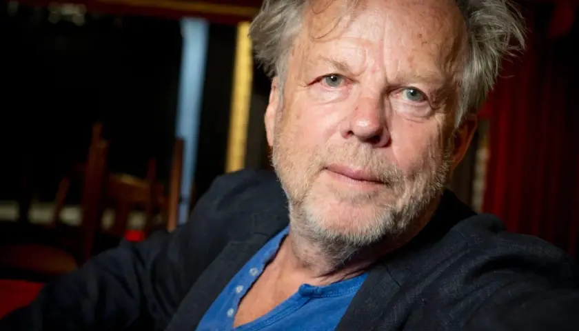 En äldre man med grått hår och skägg, klädd i kavaj och blå tröja. Han påminner om en ung Wallander.