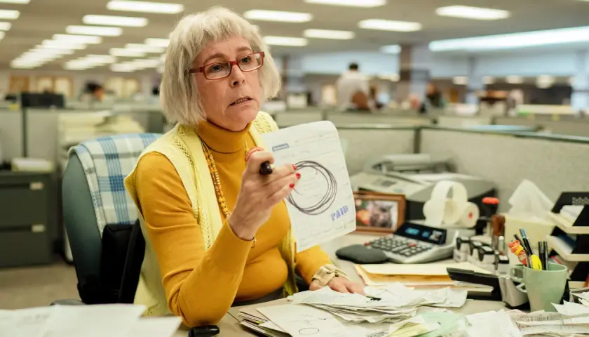 Jamie Lee Curtis som en kontorist i filmer, med glasögon och en gul polotröja, håller upp ett papper med texten "PAID".
