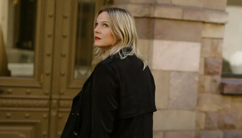 Jenny Aschenbrenner, med rött läppstift och blont hår, tittar uppåt mot en byggnad i sten.