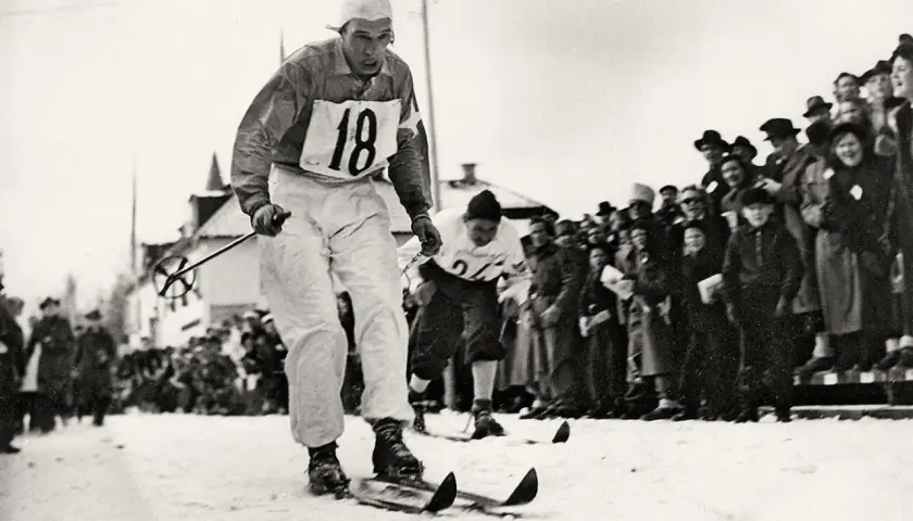 En skidåkare med nummer 18 tävlar i den vita sporten, publiken hejar.