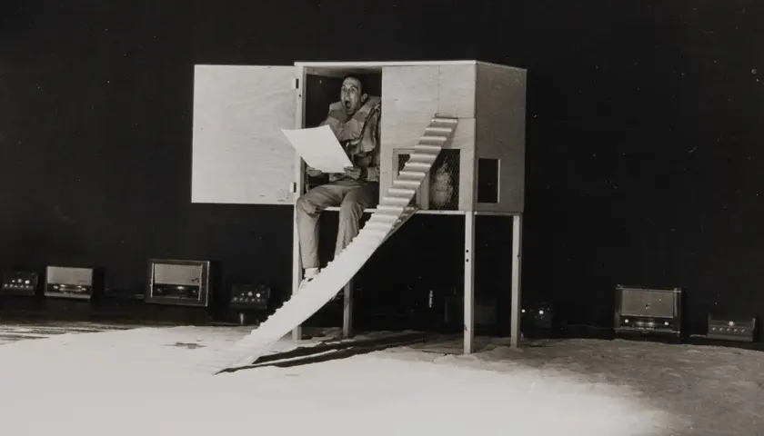 En man sitter i en träkonstruktion och läser högt ur ett papper. Robert Rauschenberg kan ha inspirerat denna scen med sina experimentella verk.