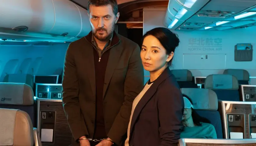 Man med handbojor och kvinna står i ett flygplan. En sovande passagerare syns i bakgrunden. Scen från konspirationen serie.