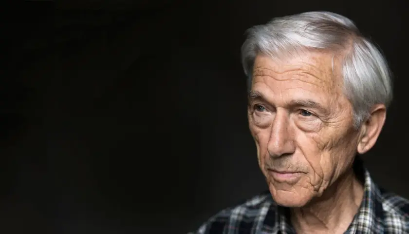 Porträtt av en äldre man med grått hår och rynkor, Theodor Kallifatides, som blickar åt sidan mot en mörk bakgrund.