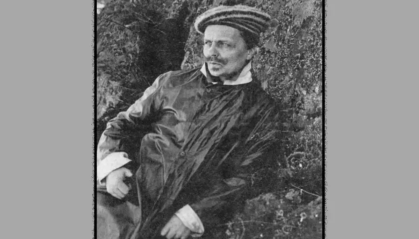 August Strindberg, klädd i rock och randig mössa, vilar mot en stenig bakgrund. Han ser ut som om han funderar på sitt nästa verk.