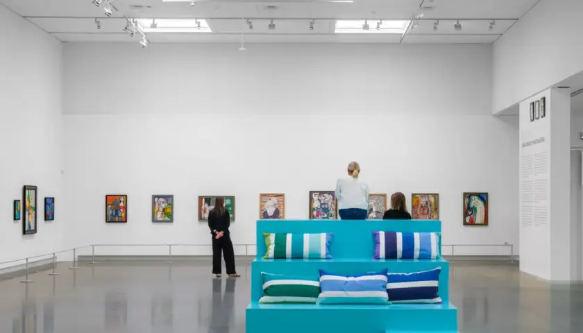I en konsthall sitter besökare på en turkos scen med kuddar, omgivna av färgglada tavlor. Kanske är de där för att inspireras, för att vara en musa för konstnären.