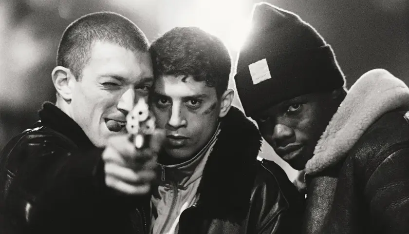 Tre unga män, en med en pistol riktad mot kameran, i en dramatisk scen från "La Haine" (Medan vi faller).