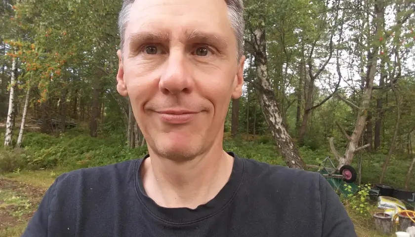 Johan Nilsson ler mot kameran i en skogsmiljö med träd och buskar.
