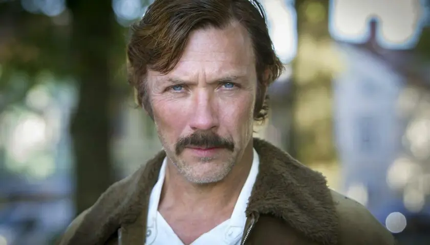 En manlig svensk skådespelare med blå ögon och mustasch, klädd i en brun jacka med foder.