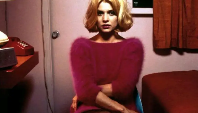 En kvinna med blont hår sitter i ett rum, kanske på ett motell. Hon bär en rosa tröja. En röd telefon syns på en hylla. Scen från "Paris, Texas".