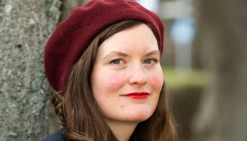 Alva Dahl, med en vinröd basker och röda läppar, ler mot kameran. Hon står utomhus vid ett träd.
