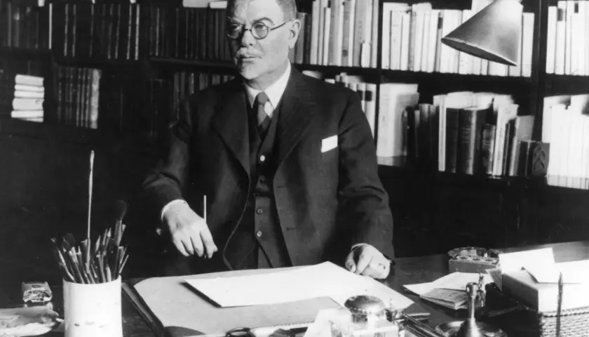 Albert Engström, med glasögon och kostym, sitter vid sitt skrivbord med böcker i bakgrunden. Penslar och bläckhorn syns på bordet.