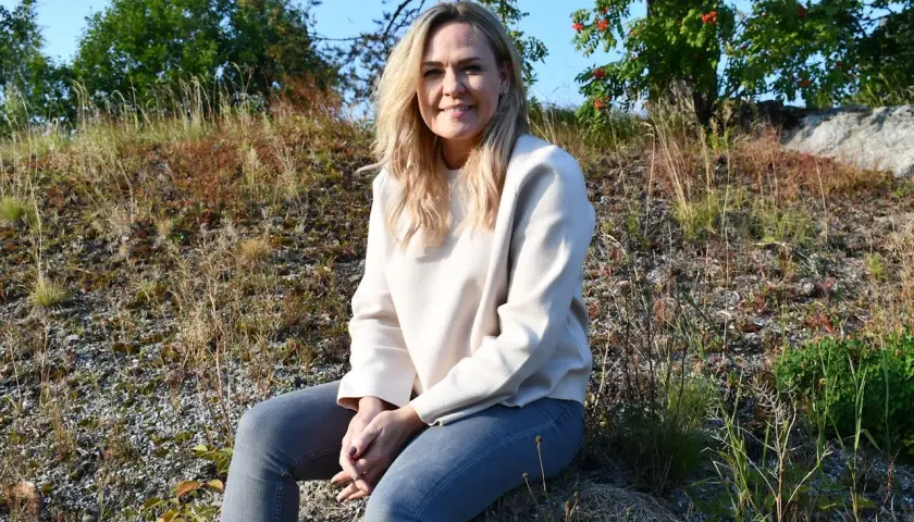 Ulrika Rolfsdotter sitter på en sten utomhus, omgiven av gräs och träd.