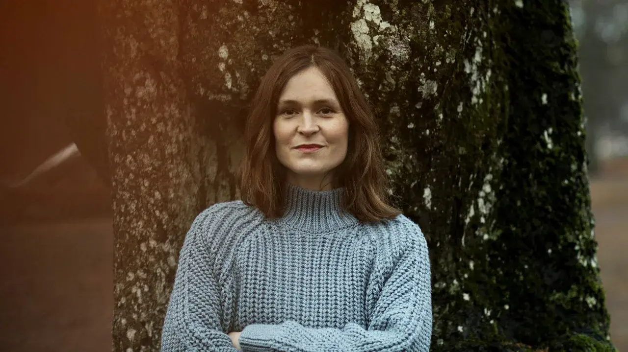 Elin Cullhed, klädd i en ljusblå stickad tröja, står med armarna i kors lutad mot ett mossigt träd.
