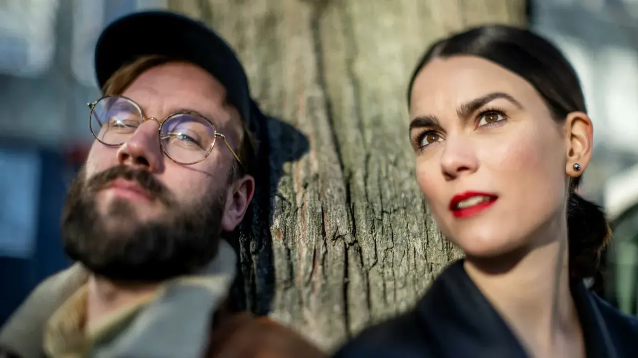 En man med glasögon och skägg lutar sig mot ett träd, bredvid sin mika partner.