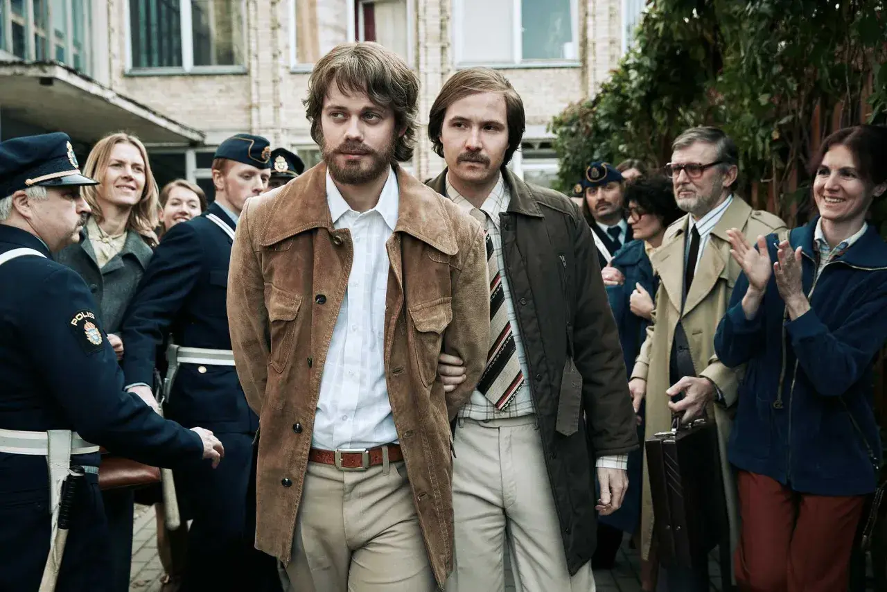 Två män förs till av poliser. En av dem, med bärnstensfärgad jacka, är Clark Olofsson i den hyllade clark olofsson serie.