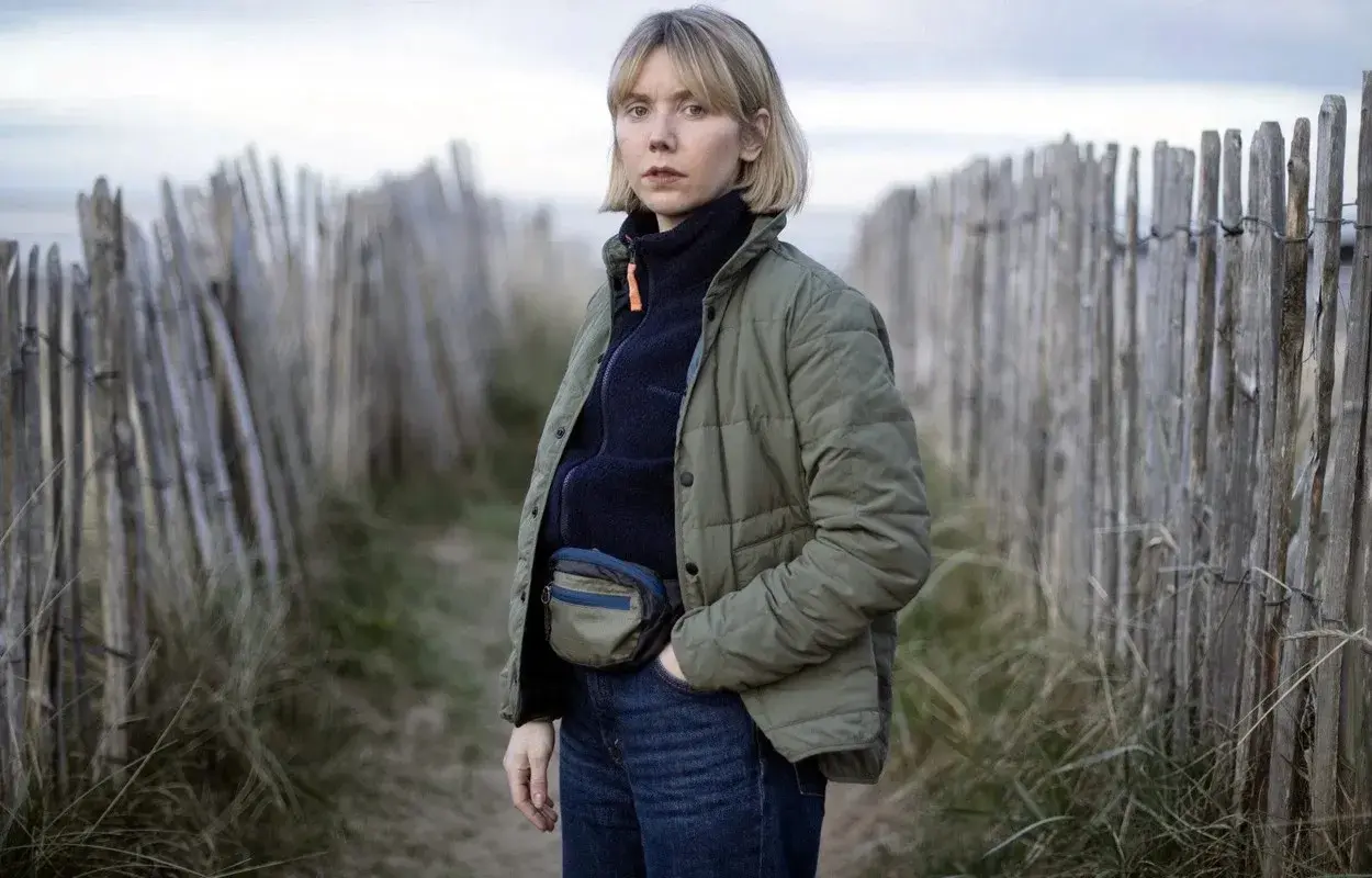 Karen Pirie står vid en vindpinad strand, klädd i en grön jacka och jeans. Hon bär en midjeväska, redo för ett nytt fall.