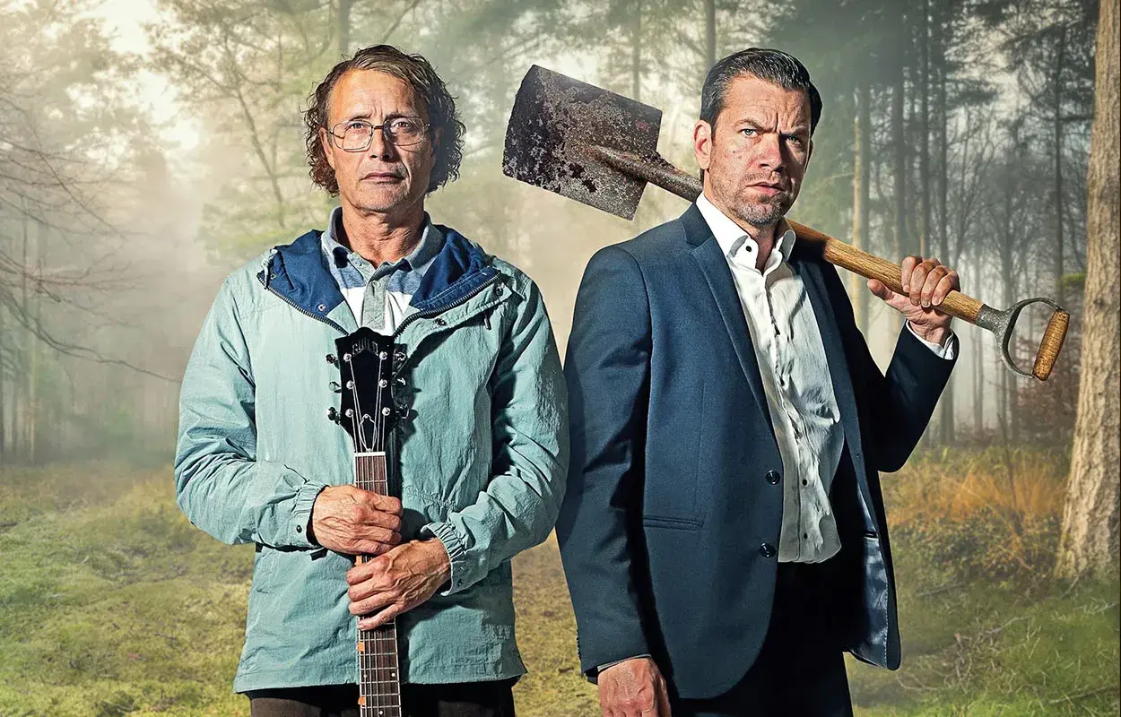 Två män i skogen, en med gitarr, en med spade. En scen från en mads mikkelsen serie.