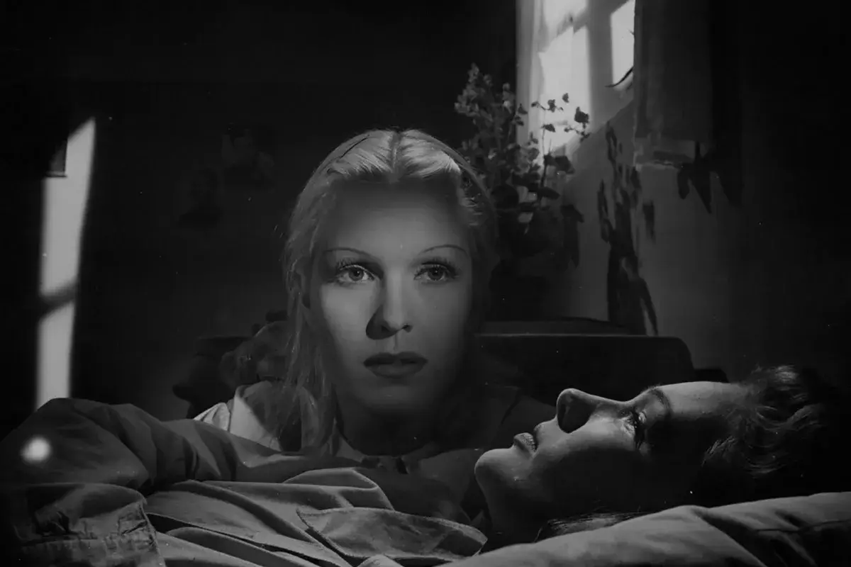 De två kvinnorna i dramatisk belysning. En kvinna ligger ner, medan den andra tittar ner på henne med intensiv blick.