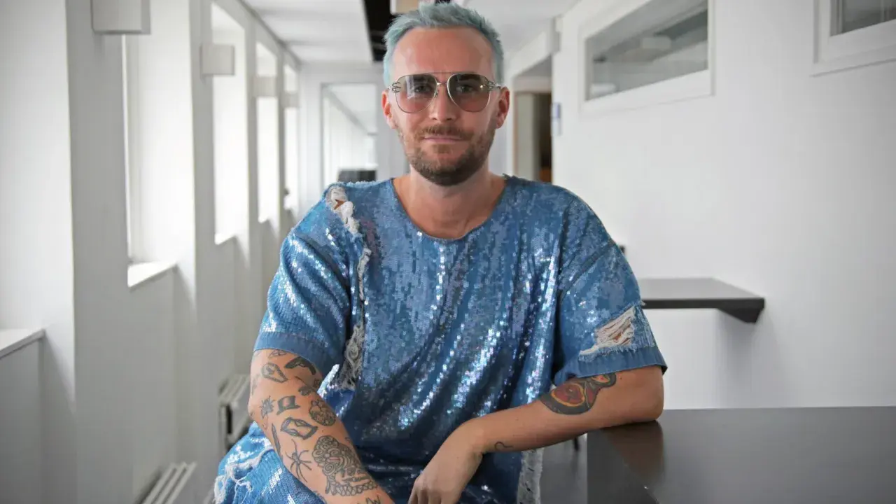 Fredrik Robertsson, med blått hår och solglasögon, sitter i en glittrig blå tröja med slitna detaljer. Hans armar är tatuerade.