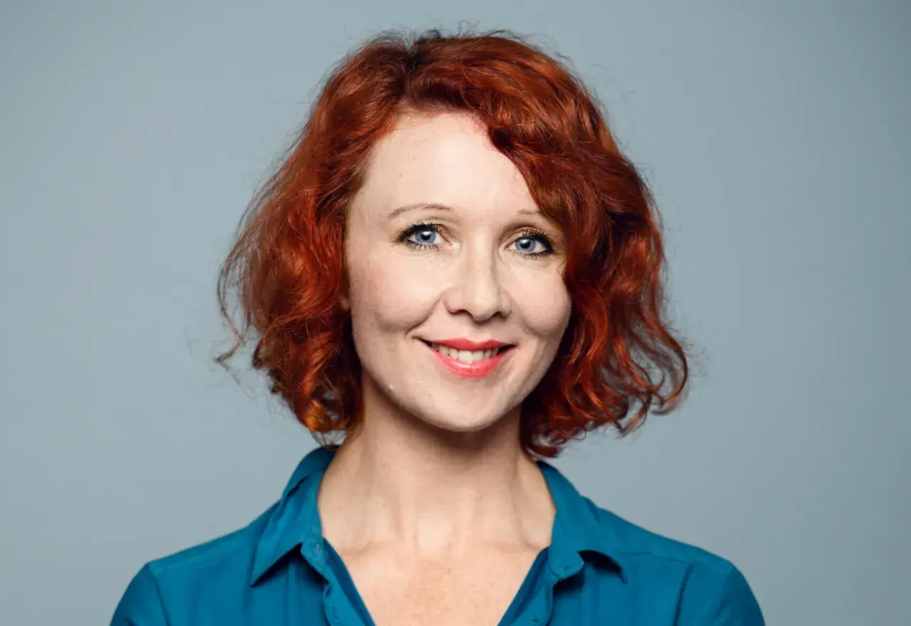 Ellinor Skagegård, en kvinna med rött lockigt hår och blå ögon, ler mot kameran i en blå skjorta.