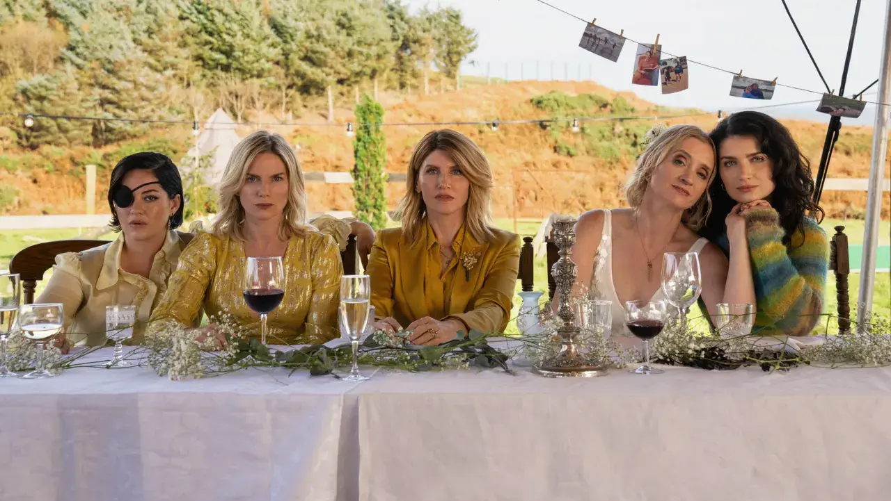 Fem kvinnor, kanske **bad sisters**, sitter vid ett dukat bord utomhus. En bär ögonlapp, en annan en glittrig gul klänning.